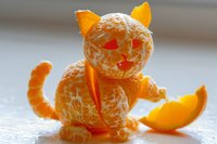 Creative food art featuring peeled mandarin orange arranged as playful cat figure with triangle peel ears, heart eyes, and curved tail on light surface.