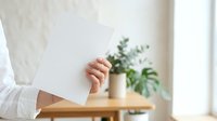 Close-up of a woman's hand holding a blank white card or paper in a bright, minimalist interior with potted green plant and wooden table.