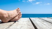 Person's bare feet resting on wooden pier overlooking calm blue ocean and sky, conveying relaxation and vacation leisure time at seaside destination.
