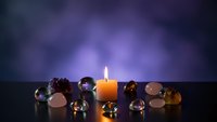 Burning candle surrounded by colorful gemstones and crystals on dark surface with purple bokeh background for meditation and spiritual practices.