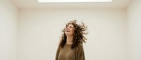 Young woman with curly brown hair dancing joyfully in minimalist white room with natural lighting from skylight overhead expressing freedom and happiness.