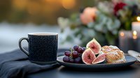Fresh figs and grapes on dark plate with pastry and black coffee cup against blurred candlelight background creating cozy autumn atmosphere.