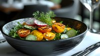 Fresh vegetable salad with roasted cherry tomatoes, cucumber, radish, and dill garnish served in black ceramic bowl on restaurant table.
