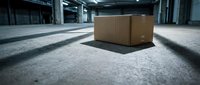 Large cardboard shipping box sitting on concrete warehouse floor with industrial lighting and shadows creating dramatic atmosphere for logistics concepts.