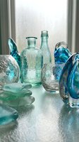 Vintage green and blue glass bottles, jars, and decorative objects arranged on reflective surface near window for home decor and interior design projects.