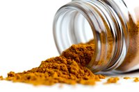 Ground turmeric spice spilling from glass jar on white surface, vibrant orange-yellow powder showing texture and natural color for cooking and wellness.