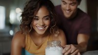 Young Hispanic woman smiling while holding glass jar in kitchen with man in background, warm lighting creates intimate atmosphere for lifestyle and relationship content.
