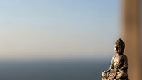 Serene Buddha statue in meditation pose against soft blurred ocean horizon background creating peaceful zen atmosphere for wellness and mindfulness concepts.
