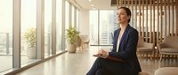 Professional businesswoman in dark suit sitting in modern office with large windows, natural lighting and contemporary interior design for corporate presentations.