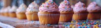 Fresh vanilla cupcakes with pink buttercream frosting and colorful sprinkles arranged in row on rustic wooden surface, selective focus on foreground.