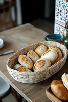 Fresh crusty bread rolls in woven basket on wooden table, natural light setting creates warm, appetizing mood for food photography and restaurant menu.