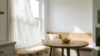 Bright minimalist dining nook with round wooden table, white cushioned bench seating, and natural light from window with sheer curtains for interior design and home decor projects.