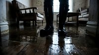 Person standing in flooded indoor space with water covering floor, wooden furniture partially submerged, emergency situation requiring cleanup and restoration.