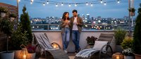 Young couple enjoying romantic evening on urban rooftop terrace with string lights, city skyline view, cozy outdoor furniture and plants for lifestyle content.