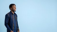 Young man with dark skin tone and braided hair wearing navy blue denim shirt, standing against light blue background, looking to side with confident expression, suitable for corporate, lifestyle, and professional website use.