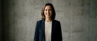 Professional businesswoman in dark blazer smiling confidently against concrete wall background. Corporate executive portrait for business presentations.