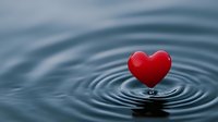 Red heart shape on rippling water surface creating concentric circles, dramatic lighting against dark blue background for love, peace and meditation concepts.
