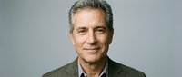 Mature man with gray hair and light complexion wearing olive green casual jacket over white shirt, smiling at camera against neutral gray background for corporate and professional use.