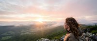 Woman with long brown hair sitting on rocky mountain peak overlooking vast green valley landscape at golden hour sunrise or sunset. Outdoor adventure and travel concept.