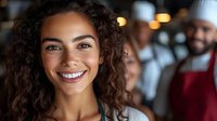 Young mixed race woman with curly hair and bright smile in restaurant setting with blurred staff in background, expressing joy and confidence.