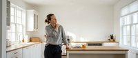 Young professional woman talking on phone in modern white kitchen with wooden countertops and natural light streaming through windows.