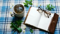 Cozy flat lay with herbal tea in ceramic mug, open leather journal, reading glasses, mint leaves and chamomile on blue plaid fabric.