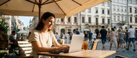 Young professional woman working on laptop at outdoor cafe terrace in historic city square during sunny day, remote work lifestyle concept.