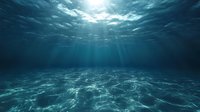 Underwater view from ocean floor with sunlight rays penetrating through turquoise water surface, creating dramatic light patterns on sandy seabed.