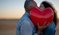 Mature couple kissing behind red heart balloon at sunset, expressing romance and love on anniversary or Valentine's Day celebration.