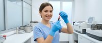 Female laboratory technician in blue scrubs and gloves holding test tube in modern medical research facility for scientific analysis and healthcare testing.