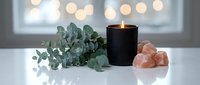 Spa wellness setup with eucalyptus branch, lit black candle and pink himalayan salt crystals on white surface creating relaxing aromatherapy atmosphere.