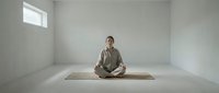 Young Asian woman practicing meditation in minimalist white room with natural light streaming through window, sitting cross-legged on yoga mat in peaceful pose.