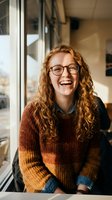 Happy young Caucasian woman with curly red hair and glasses laughing in modern office space wearing cozy knit sweater for workplace positivity concepts.