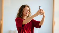Happy young Caucasian woman with curly hair taking selfie with smartphone, smiling joyfully in red sweater against warm indoor lighting background.