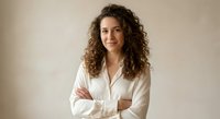 Professional young woman with curly hair in white shirt smiling confidently with crossed arms against neutral background for business presentations.
