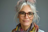 Confident mature woman with silver hair and stylish glasses smiling warmly at camera, wearing colorful scarf against gray background. Professional portrait.