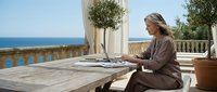 Young blonde woman in beige robe working on laptop at luxury Mediterranean terrace with sea view, remote work lifestyle concept for digital nomads.