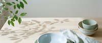 Minimalist tabletop arrangement with stacked ceramic bowls, white plate, linen napkin, and green foliage casting natural shadows on neutral surface for home decor and lifestyle styling.