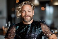 Caucasian man with blonde hair and beard wearing black sequin sheer top smiles warmly at camera. Blurred restaurant background creates soft bokeh effect.