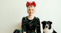 Woman with blonde hair wearing red bow headpiece and dark green velvet top with gold details, posing with black and white border collie dog against neutral background for holiday or festive portraits.
