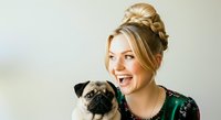 Young woman with blonde hair styled in an updo, fair complexion, wearing a dark green top, smiling while holding a pug dog with fawn and black coloring against neutral background.
