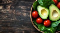 Fresh halved avocado, cherry tomatoes and baby spinach leaves in wooden bowl on dark rustic table, top view with copy space for healthy eating concept.