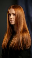 Young Caucasian woman with long glossy auburn hair posing against dark background, showcasing healthy shiny straight hairstyle for beauty and salon concepts.