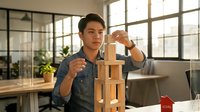 Young man with East Asian ethnicity, wearing casual denim shirt, stacking wooden blocks in modern office workspace with large windows and plants, demonstrating construction, planning, and creative problem-solving concepts.