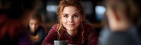 Young woman with auburn hair and warm smile holding coffee cup in cafe setting, soft natural lighting creates intimate atmosphere for lifestyle and hospitality content.