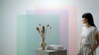 Woman with dark hair and light skin tone, wearing casual beige and white clothing, observing dried flowers in vase on table with stacked books against colorful pastel backdrop for interior design and lifestyle content.