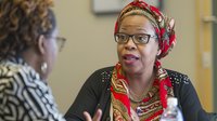African woman wearing bright red headscarf and glasses engaged in animated business conversation with colleague during office meeting, displaying positive professional interaction.