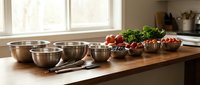 Stainless steel mixing bowls arranged on wooden kitchen counter with fresh vegetables and herbs by bright window for cooking preparation.