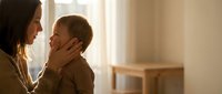 Young Asian mother tenderly holding toddler son's face in warm sunlit living room, expressing maternal love and bonding moment between parent and child.