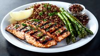 Grilled salmon fillet with charred marks served alongside quinoa, fresh asparagus, and garnished with herbs and lemon wedge on white plate, healthy meal concept.